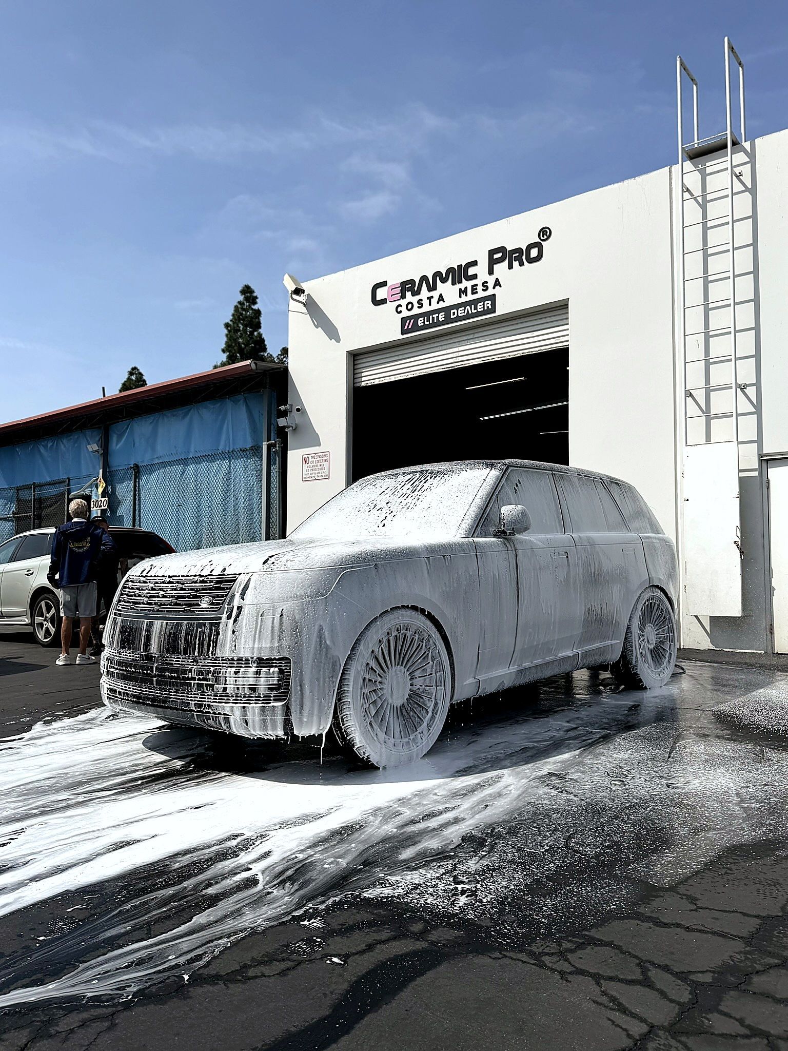 White car covered in foam at a car wash, with another vehicle and person nearby.
