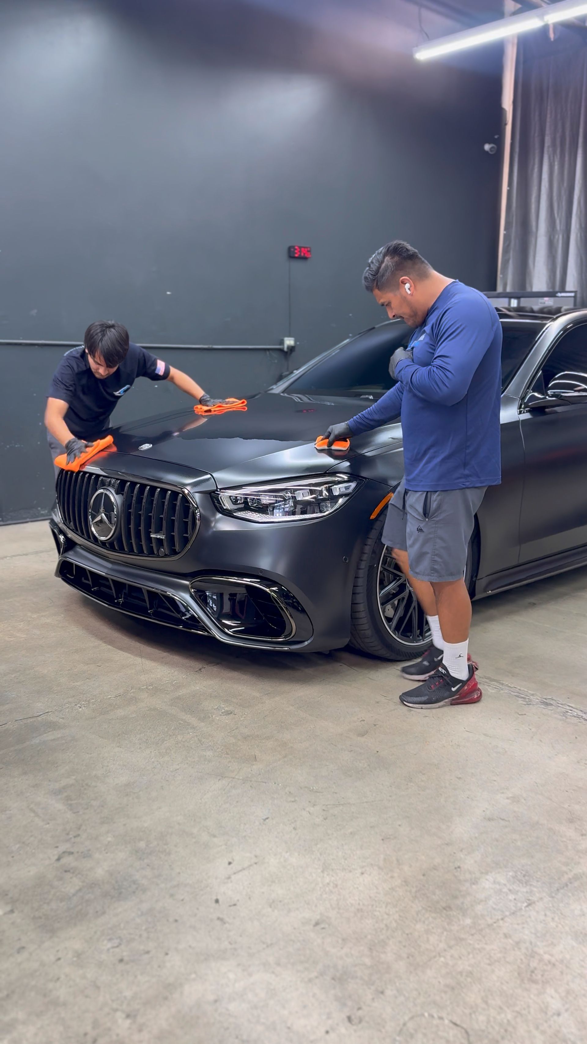Two men work on the front of a black car in a garage, one polishing the hood and one inspecting the grille.