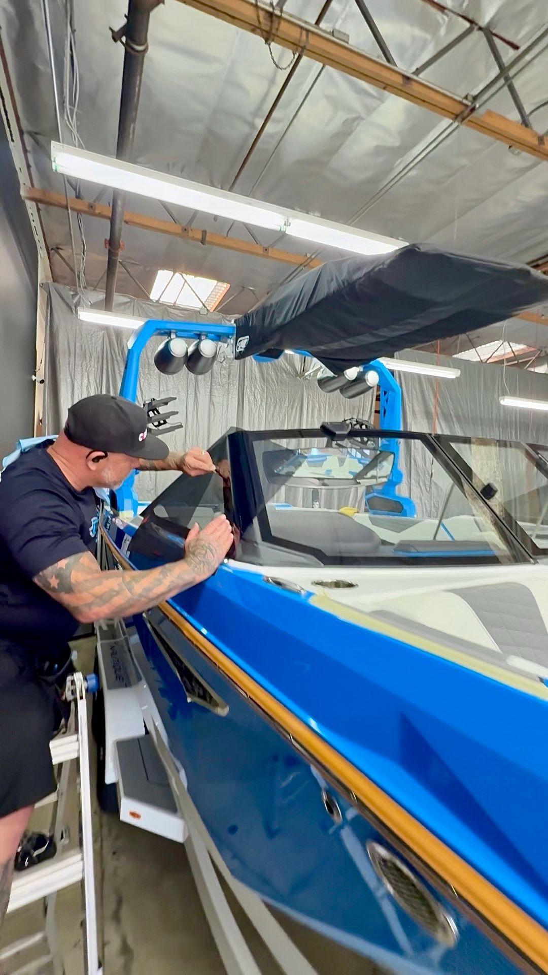 Man spraying blue boat hull in a workshop, with a masked worker leaning over the side and masking tape visible.