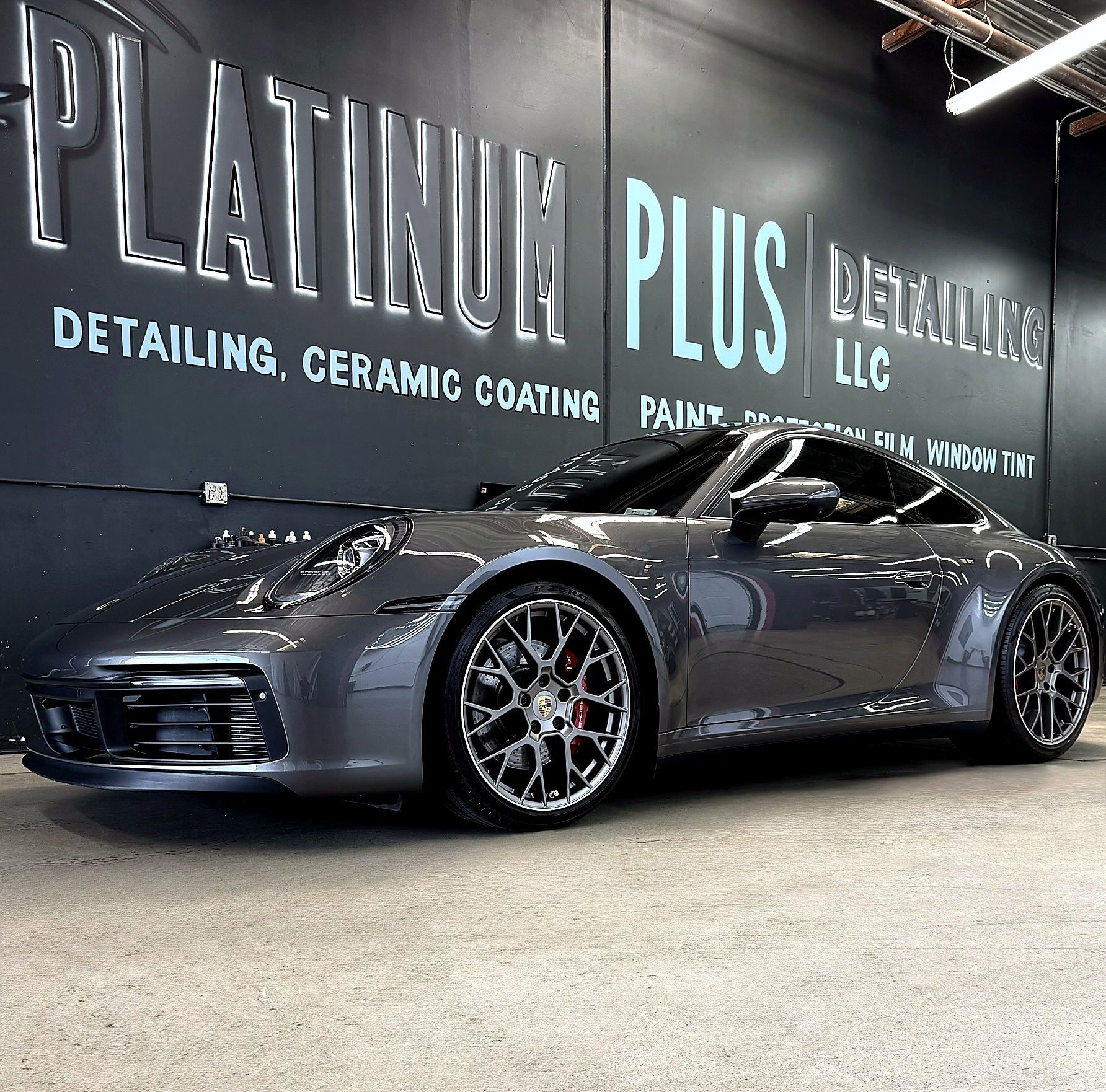 Gray sports car in a detailing shop with a black wall sign reading “Platinum Plus Detailing LLC”