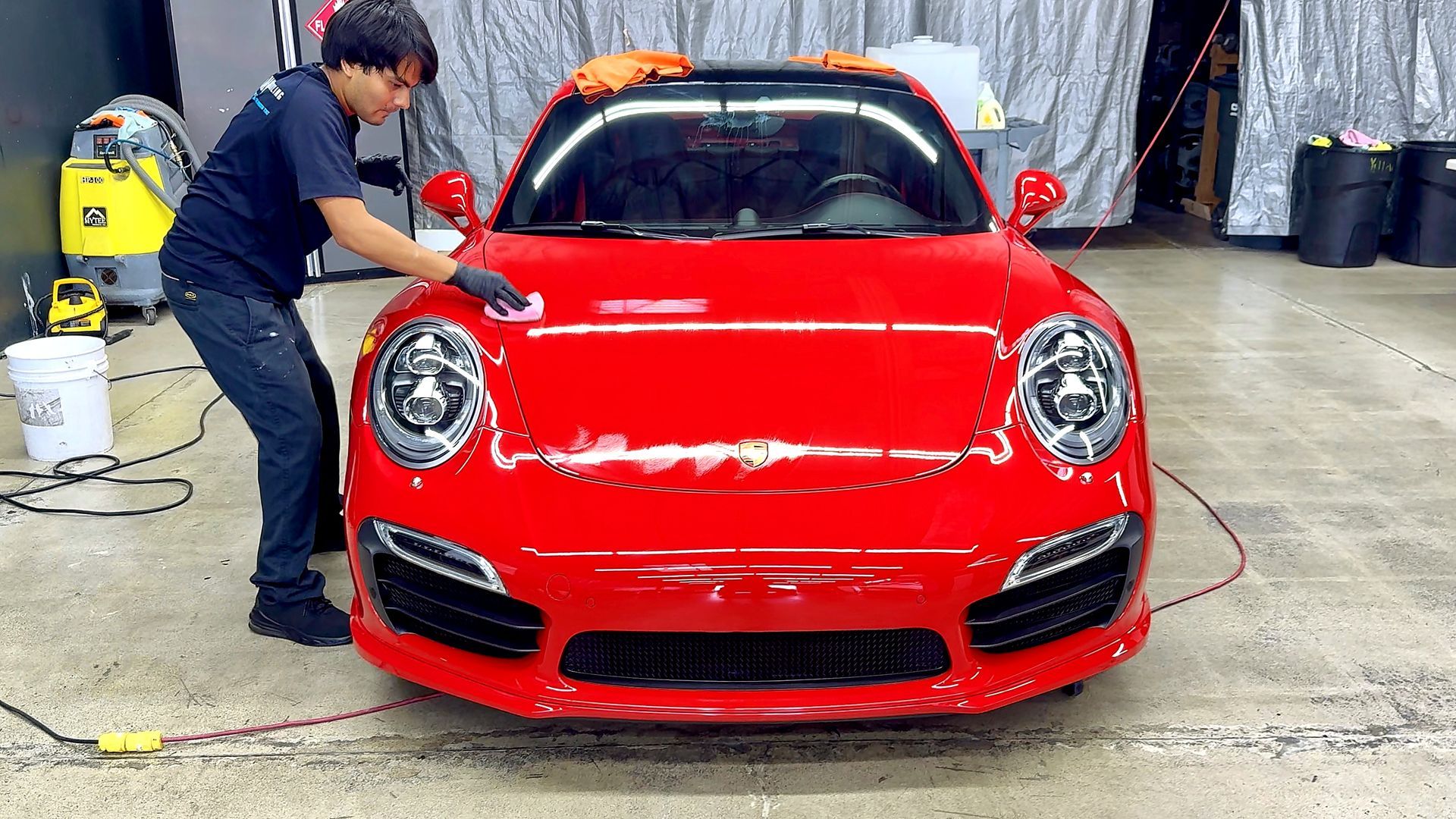 Worker detailing a red sports car in a garage, with cleaning equipment nearby.