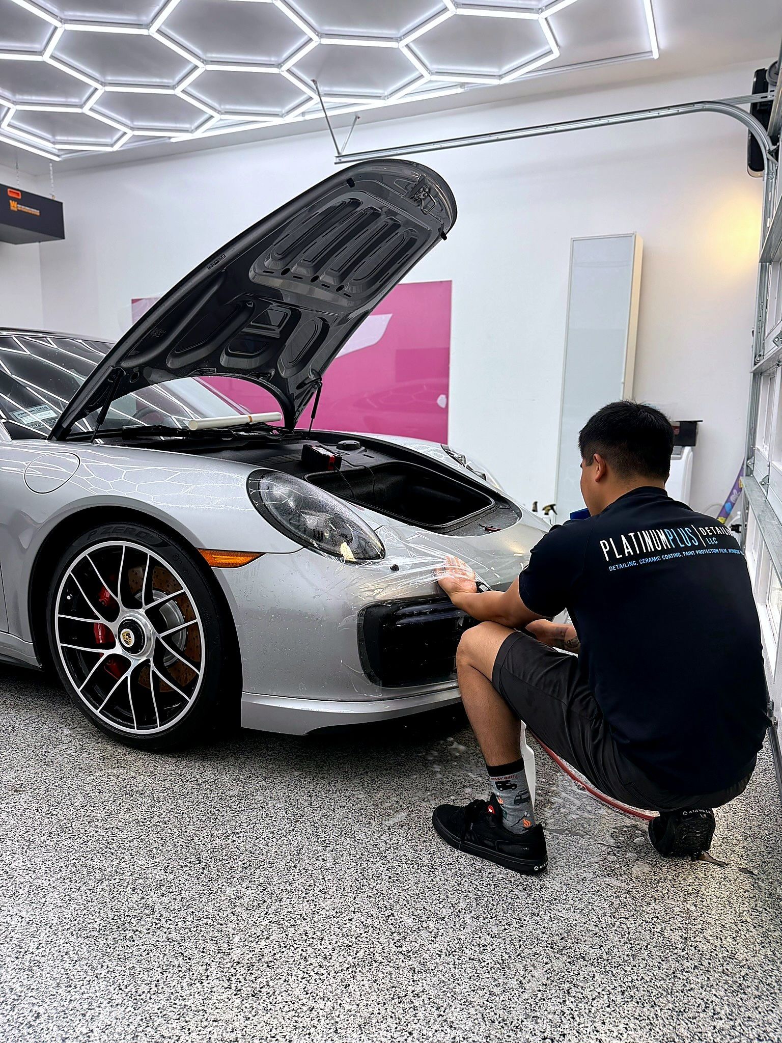 Man in a black shirt detailing the front of a silver sports car with the hood open in a bright garage