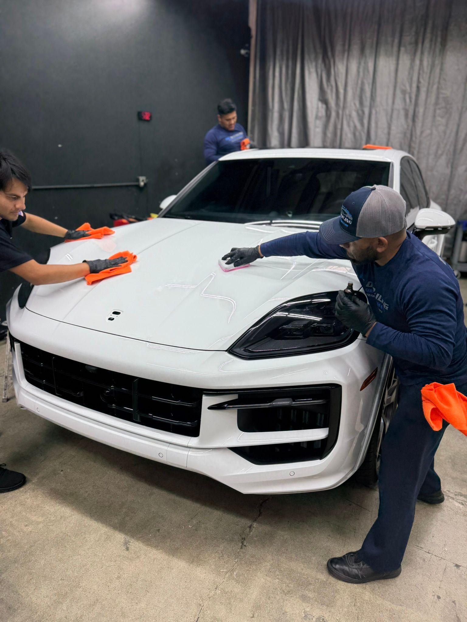 Three people wiping a white sports car indoors with orange cloths