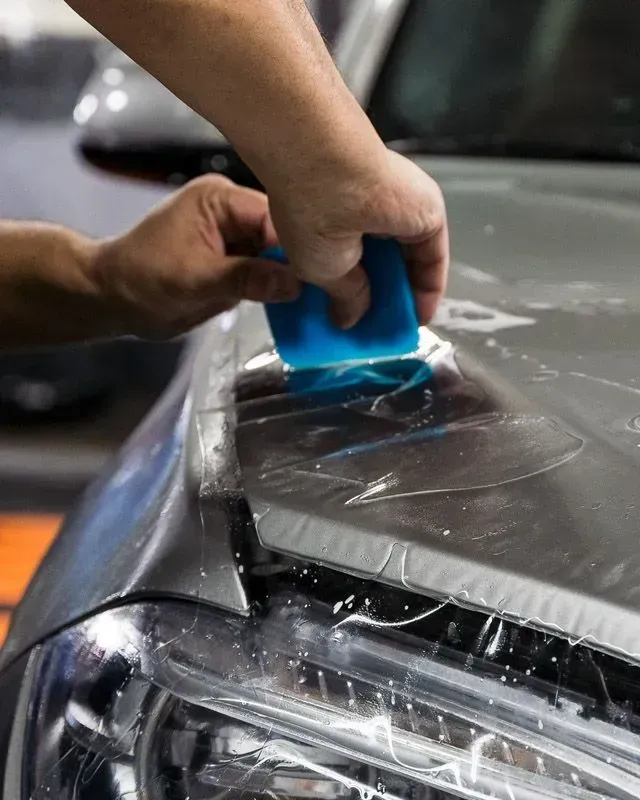 Hands applying a blue squeegee to a car hood covered in protective film, near the headlight