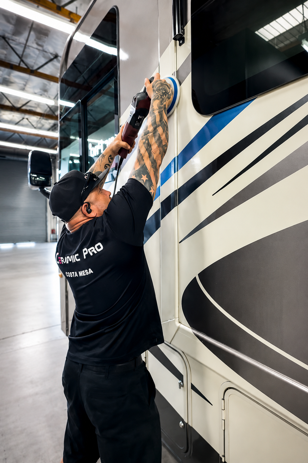 Man in black shirt washes a large white RV with blue stripes in a garage
