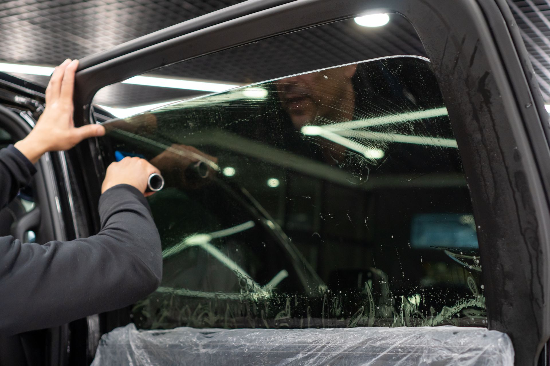 Worker installing a car window tint on a black vehicle inside a shop