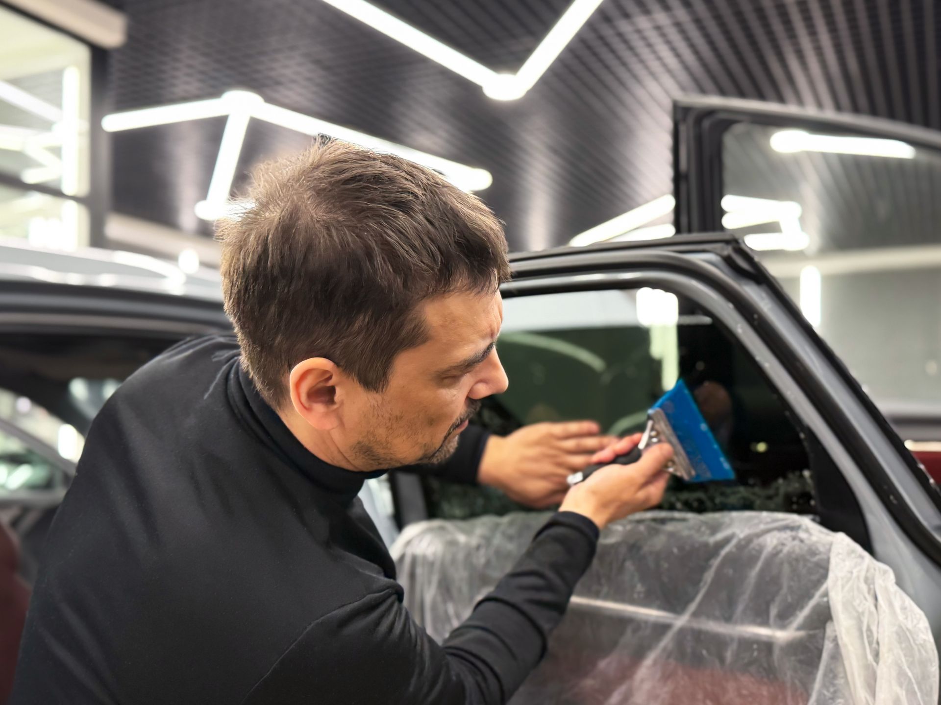 Man using a blue tool to inspect a car interior in a garage, with plastic seat covers visible.