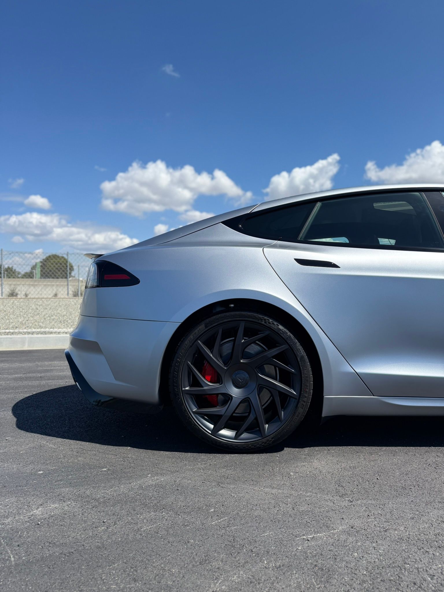 Silver sedan parked on pavement under a blue sky, showing the rear wheel and side profile.