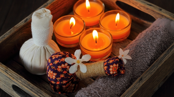 Lit orange candles in a wooden tray with decorative flowers, fabric bundles, and a towel.