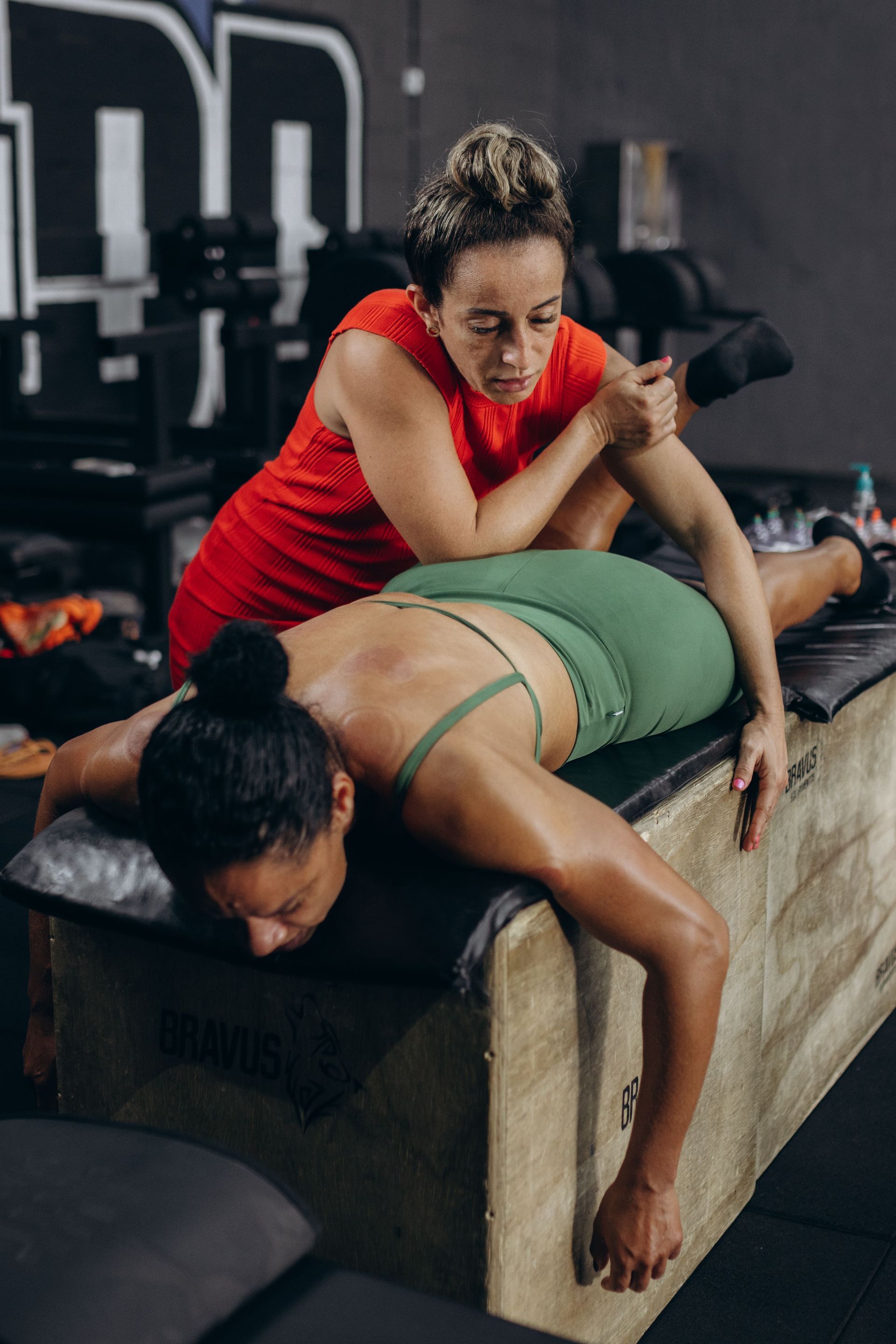 Trainer stretching a woman in green workout clothes on a gym table as she leans forward during a session