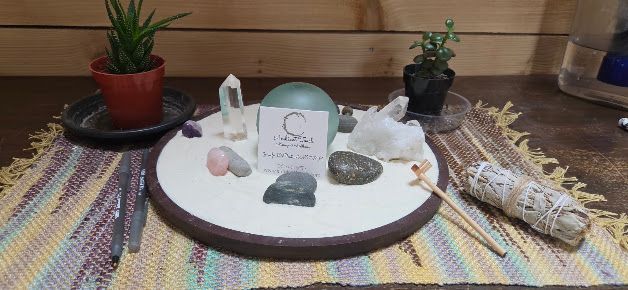 Small altar display on a table with crystals, incense, candles, and potted plants on a woven cloth.