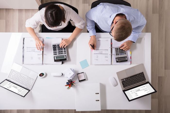 A man and a woman are sitting at a desk with calculators and laptops.