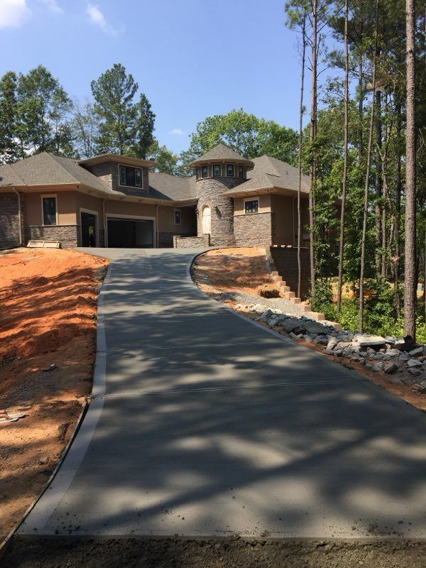A newly paved concrete driveway leads toward a tan-colored house with a stone tower, surrounded by trees and dirt.