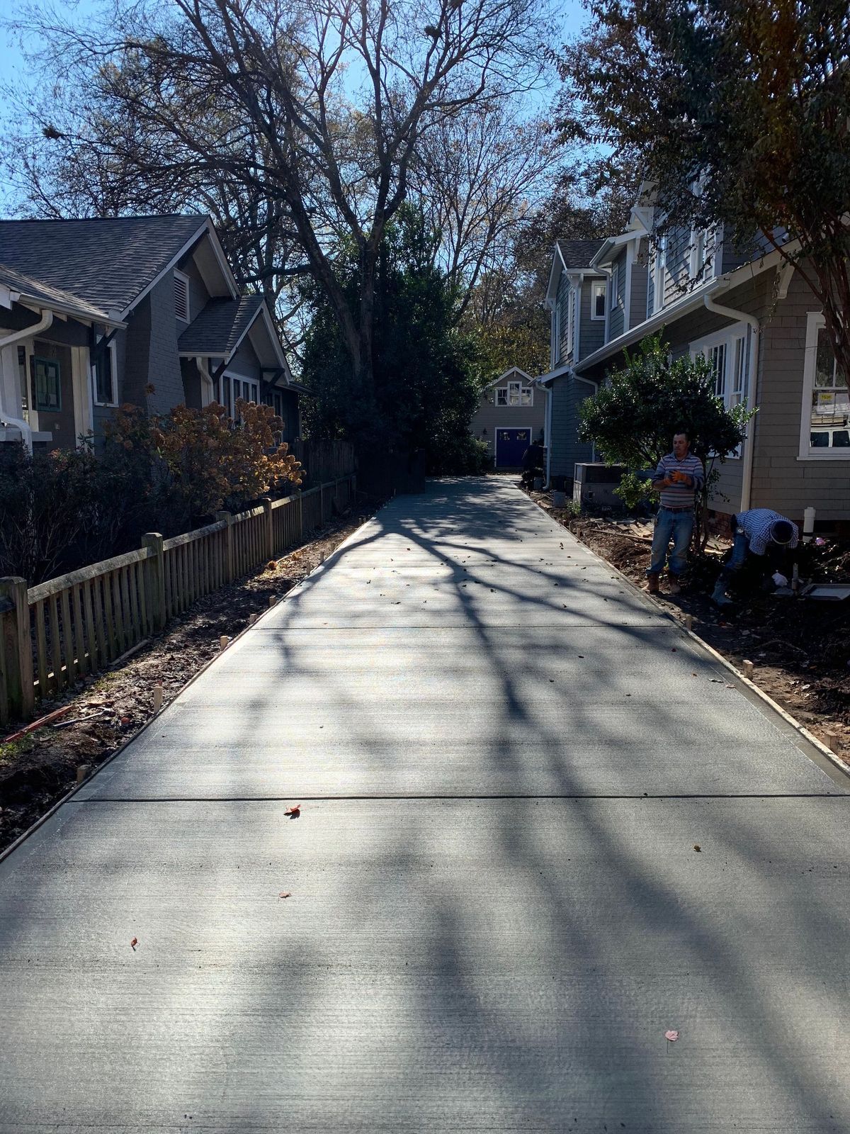 A newly poured concrete driveway leads between two houses on a sunny day, with two people working along the side.