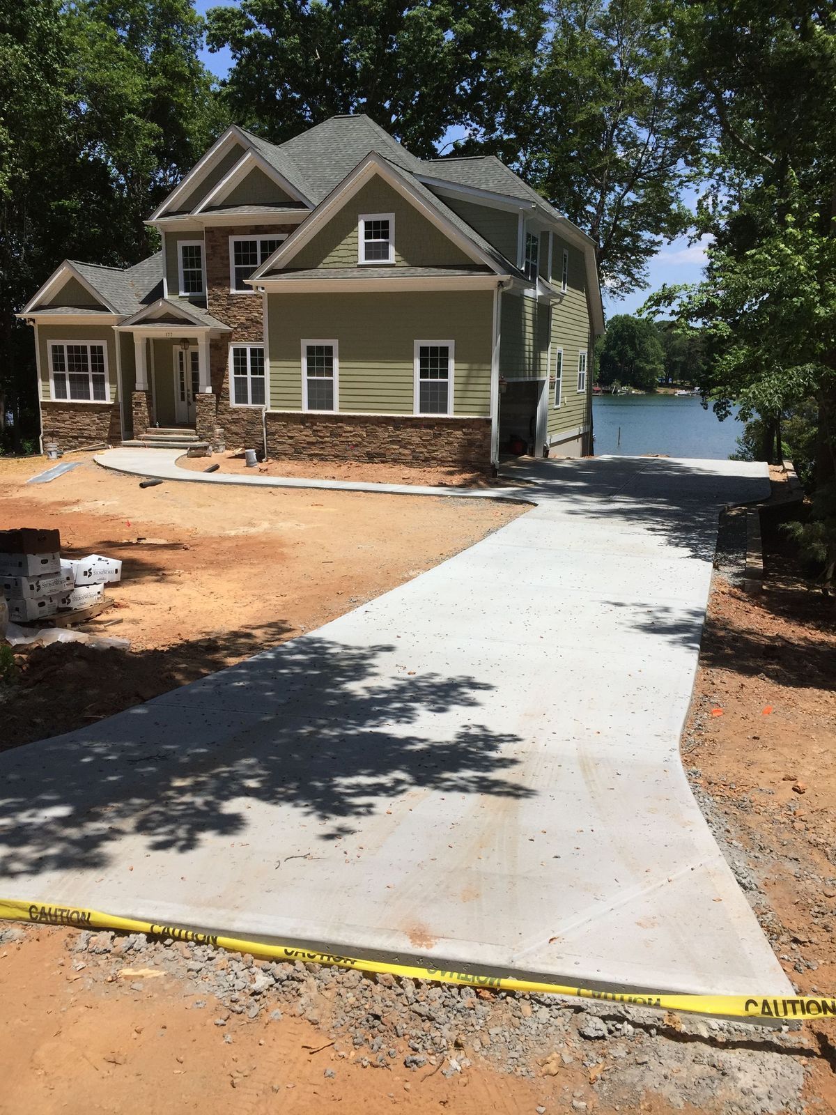 A newly poured concrete driveway leads to a modern, two-story house with green siding and stone accents near a lake.