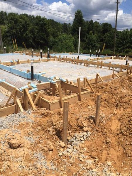 A construction site showing a concrete foundation under development with wooden framing and exposed plumbing pipes.