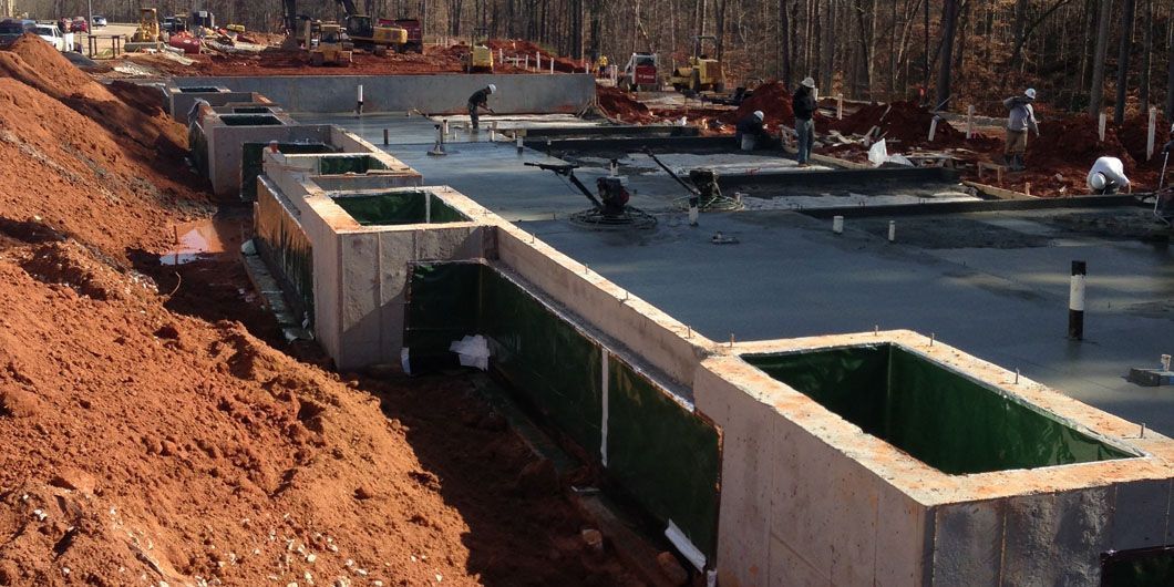 Construction workers smooth a large concrete foundation slab at a building site with exposed dirt embankments.