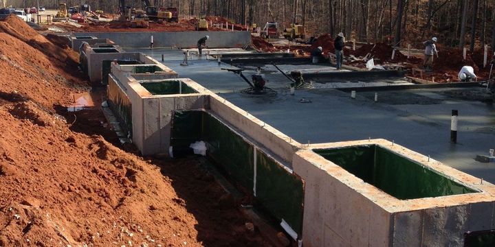 Construction workers finish a poured concrete slab foundation near a large red-dirt embankment.