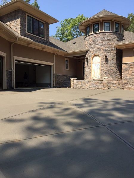 A multi-story home with a stone-clad turret, tan stucco walls, and a large concrete driveway under a clear blue sky.