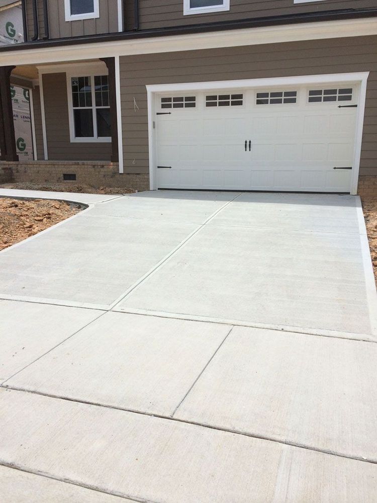 A newly poured concrete driveway leads to a closed white garage door of a brown, multi-story house under construction.