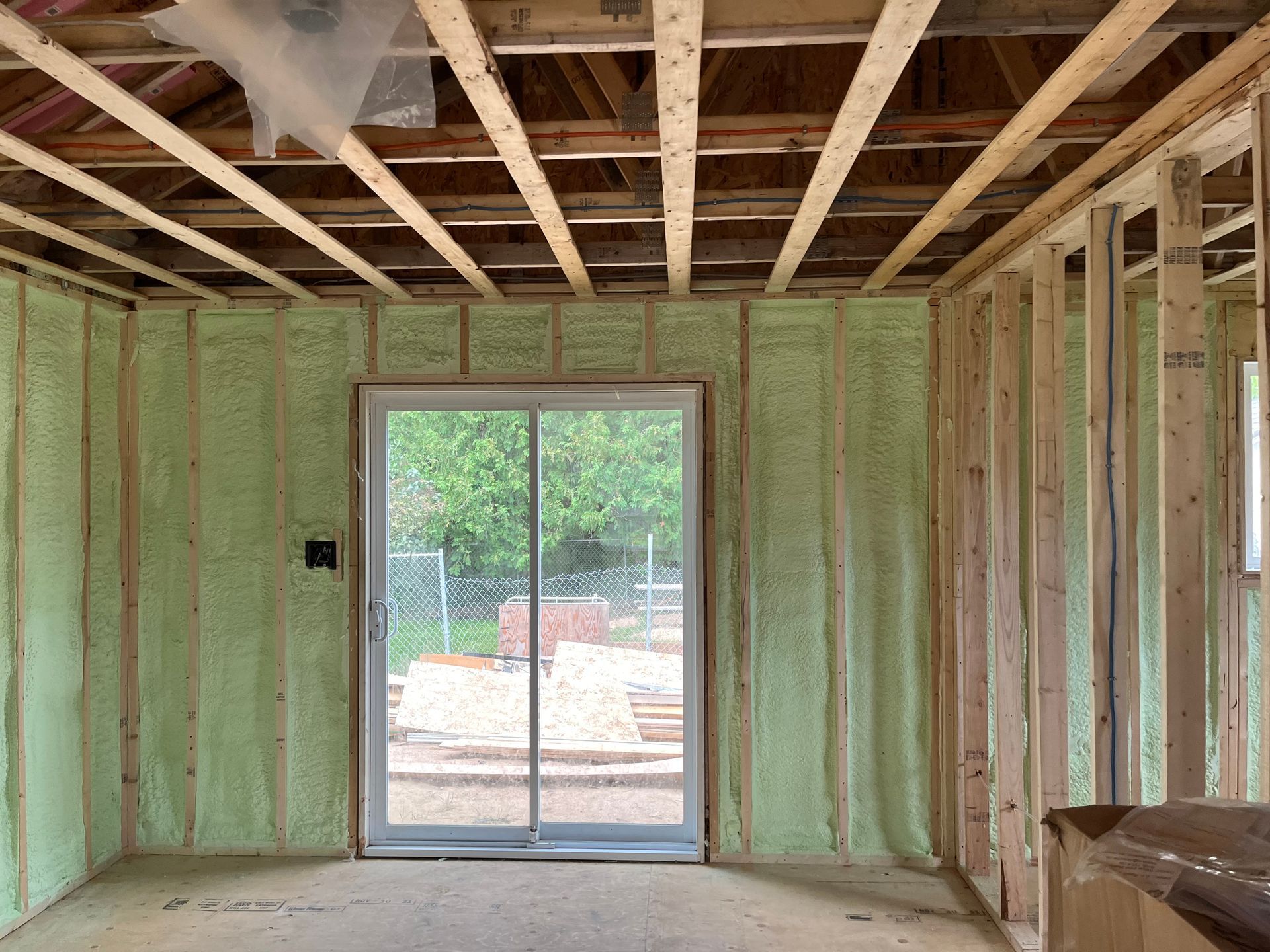 Interior view of a room under construction with green insulation, exposed beams, and a sliding glass door.