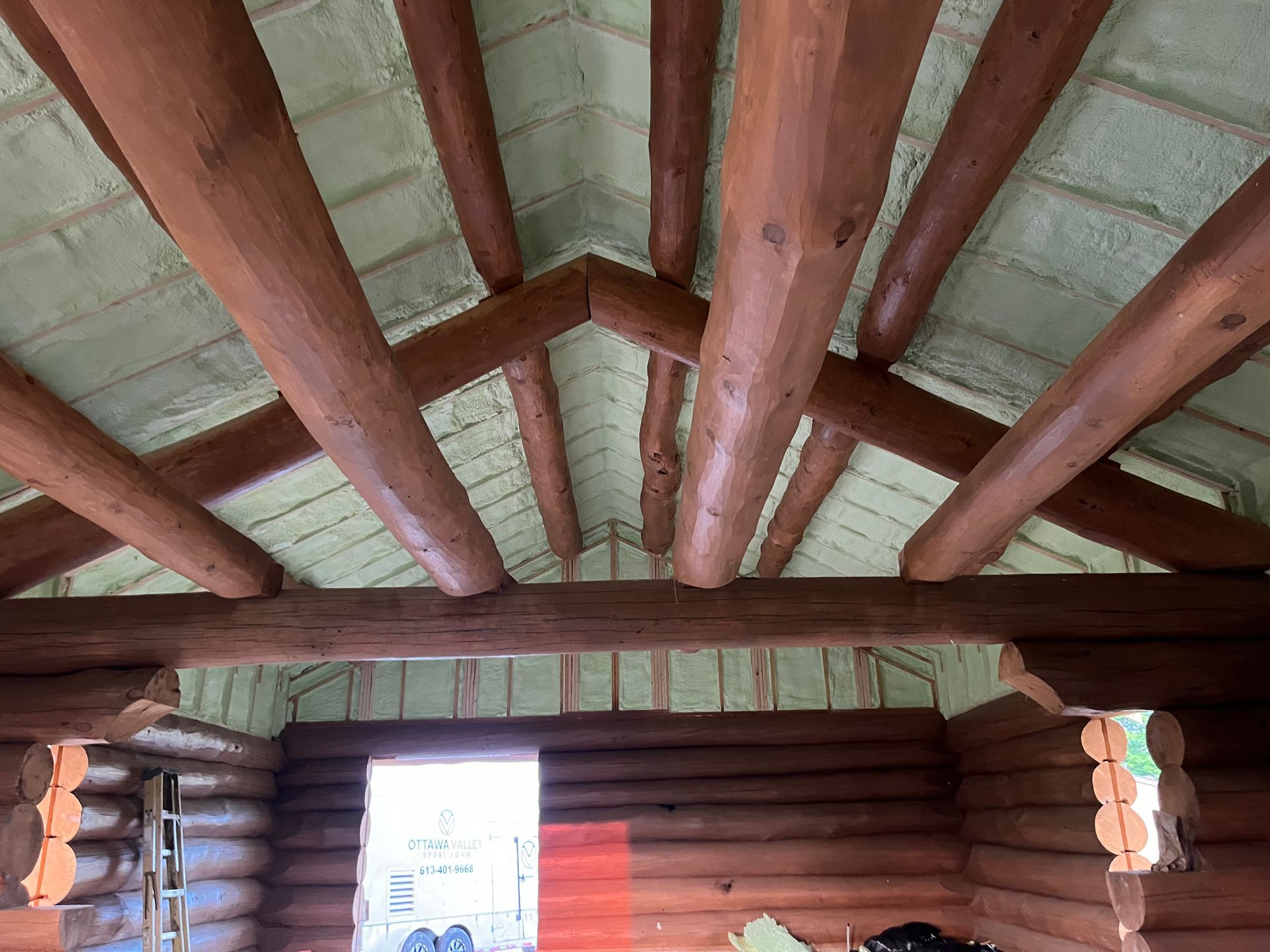 Interior of a log cabin with exposed wooden beams, spray foam insulation, and an open doorway.