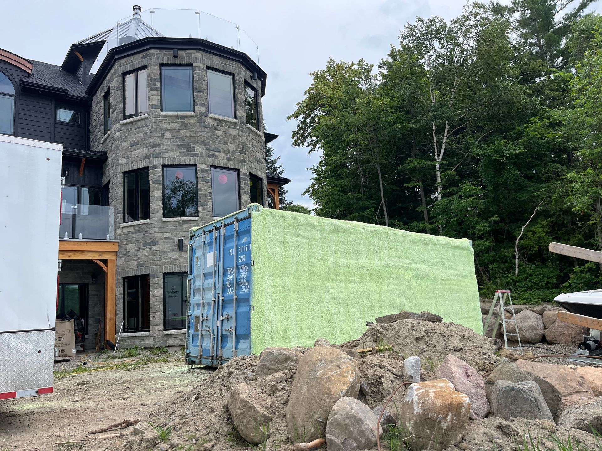 Blue shipping container covered in green foam insulation near a large stone house.