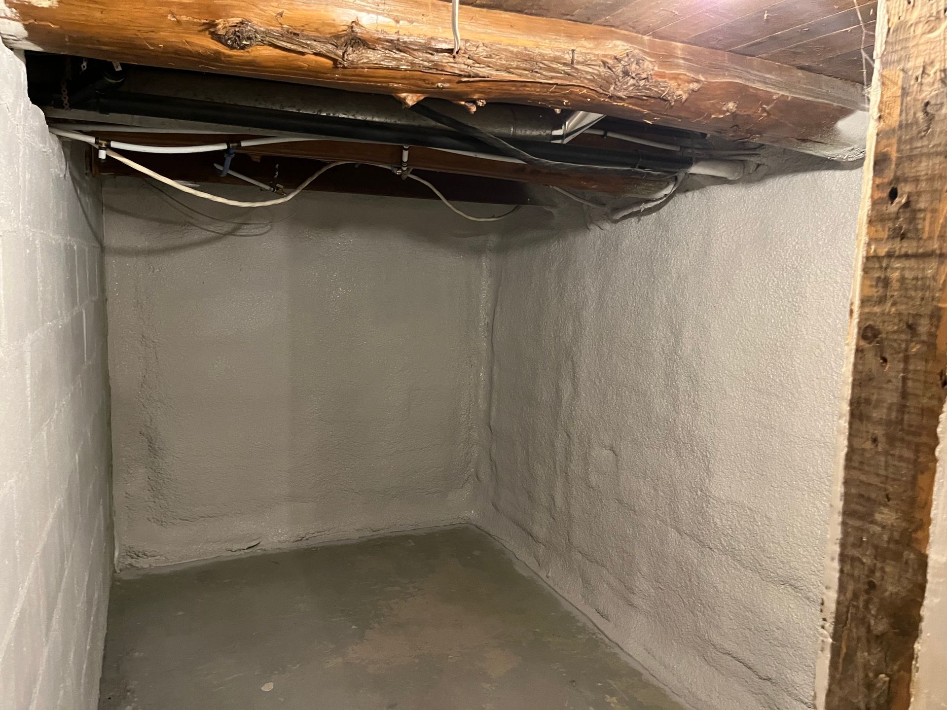 Concrete-walled basement corner, gray walls and floor, wooden beam ceiling with wires.