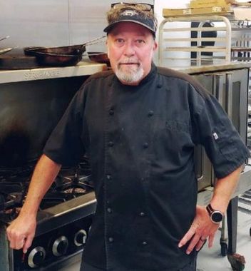 Chef in black uniform standing in a commercial kitchen beside a stove, wearing a cap and watch.
