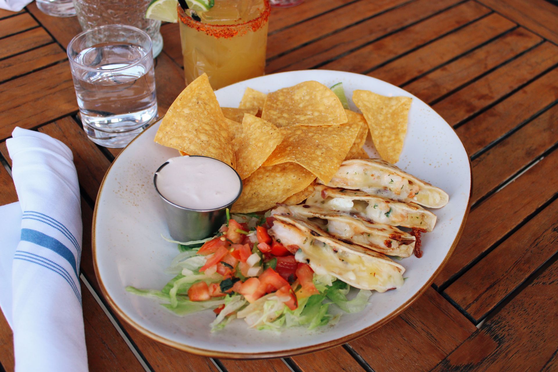 Plate of grilled chicken, chips, and fresh salsa on a wooden table with a glass of water and drink.