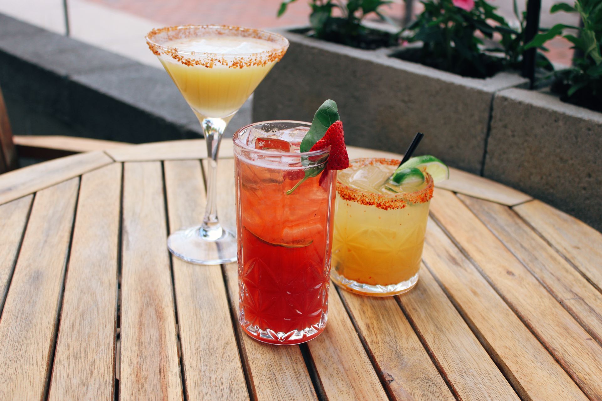 Three colorful cocktails in martini and rocks glasses on a wooden patio table.