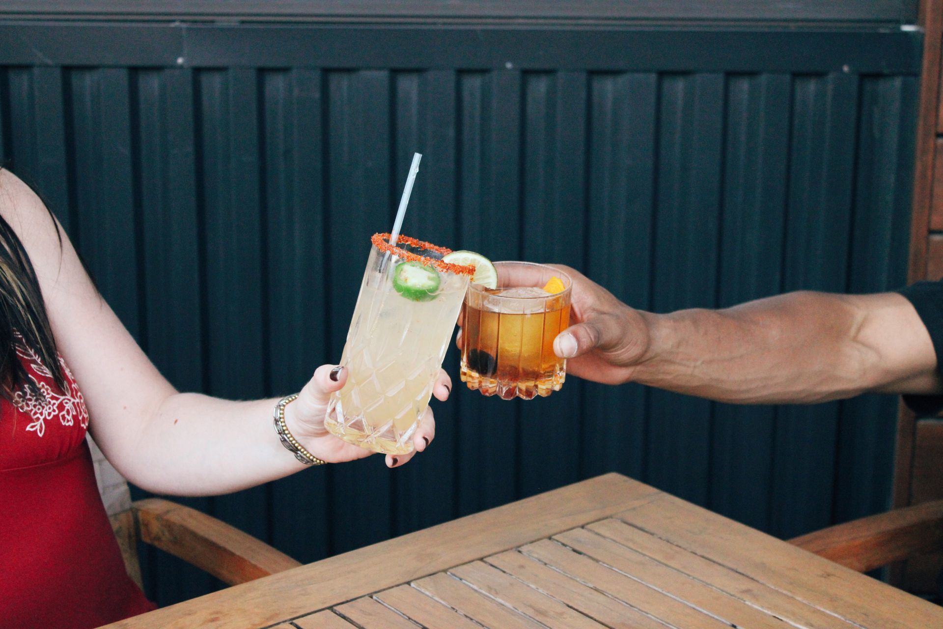 Two people clink a pale drink and an amber cocktail over a wooden table at an outdoor bar.
