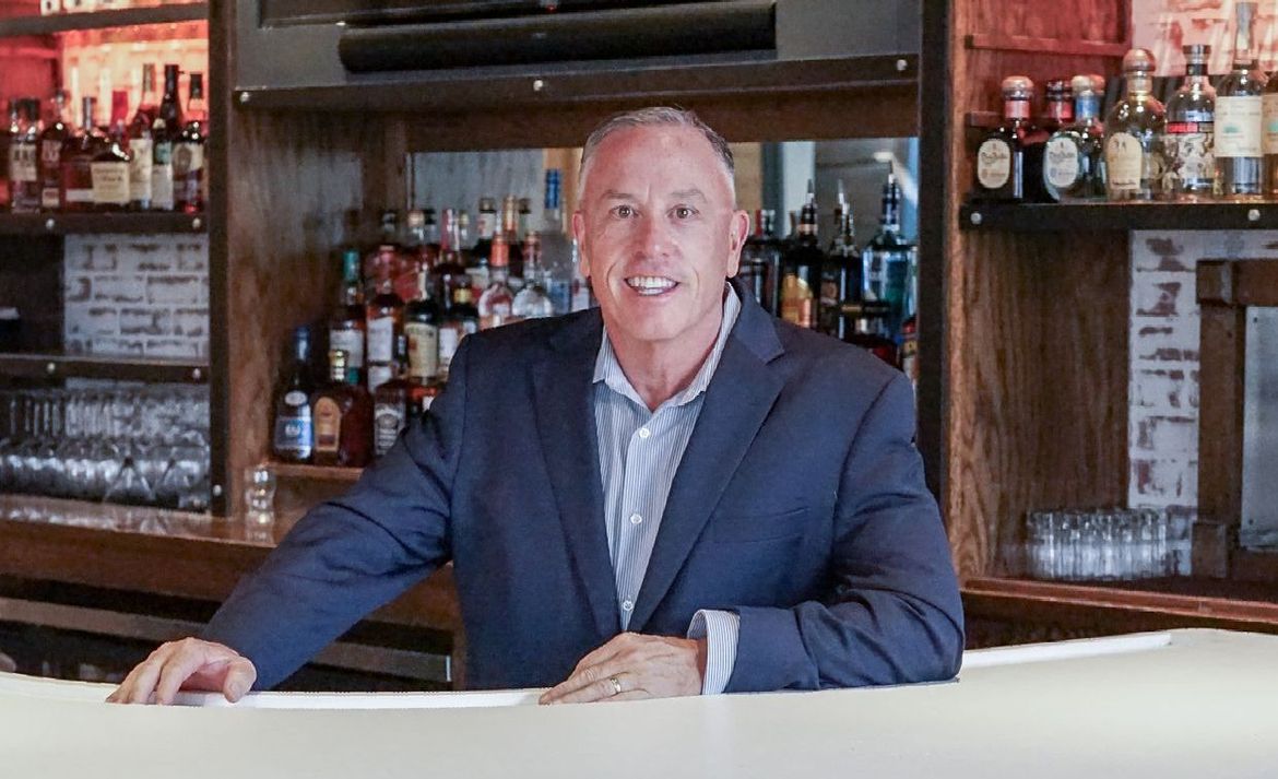 Man in a blue blazer standing behind a bar with bottles on shelves, smiling at the camera.