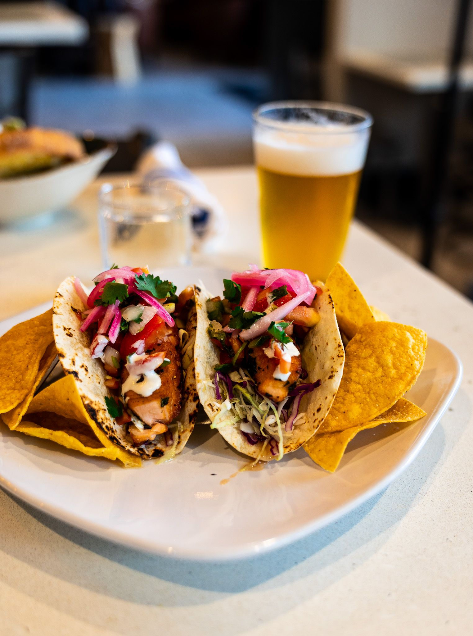 Tacos on a white plate with a glass of beer in a restaurant setting