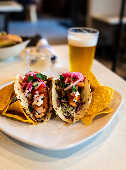 Tacos on a white plate with a glass of beer in a restaurant setting