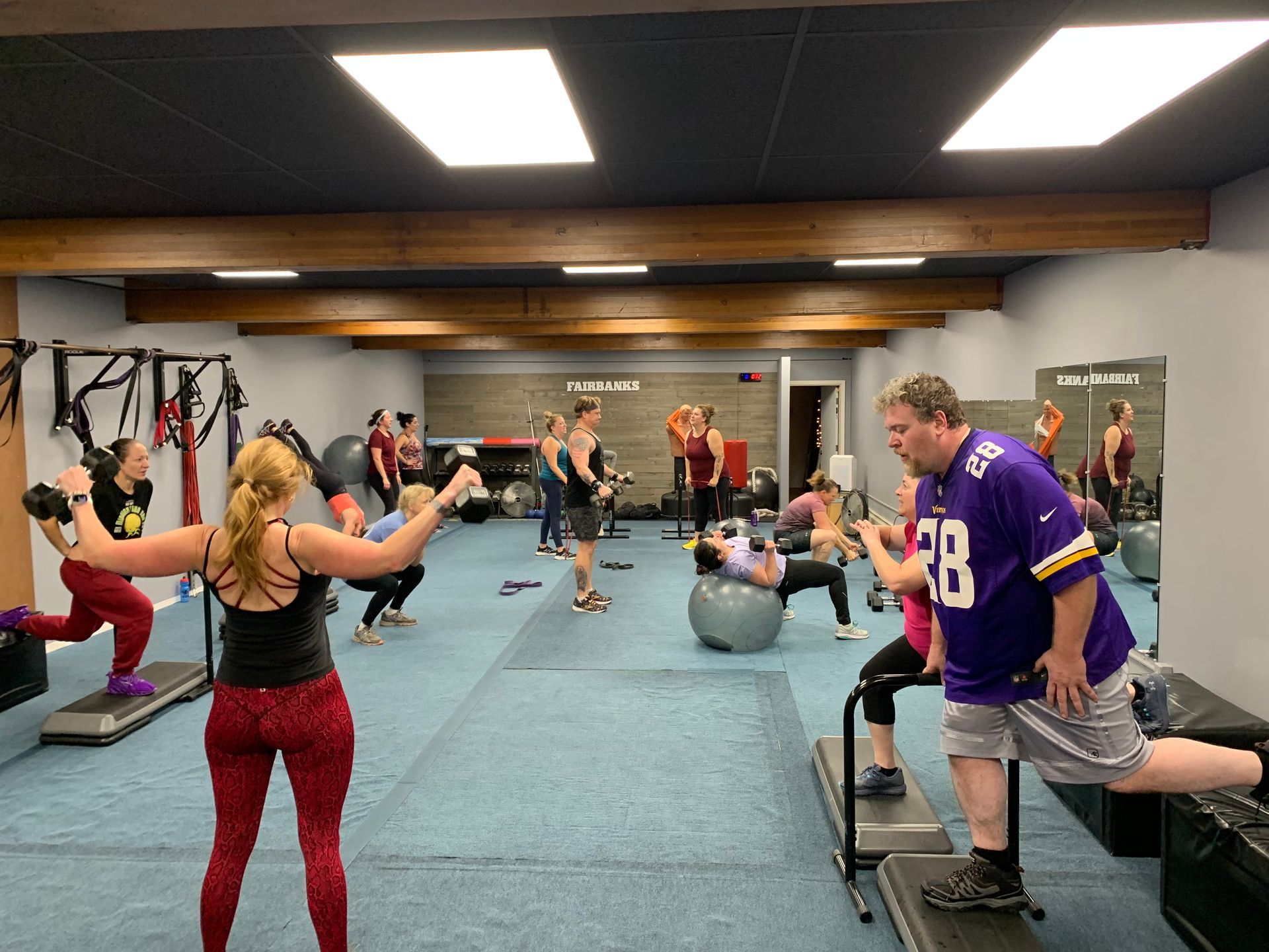 People exercising in a gym, including using dumbbells, exercise balls, and step platforms.