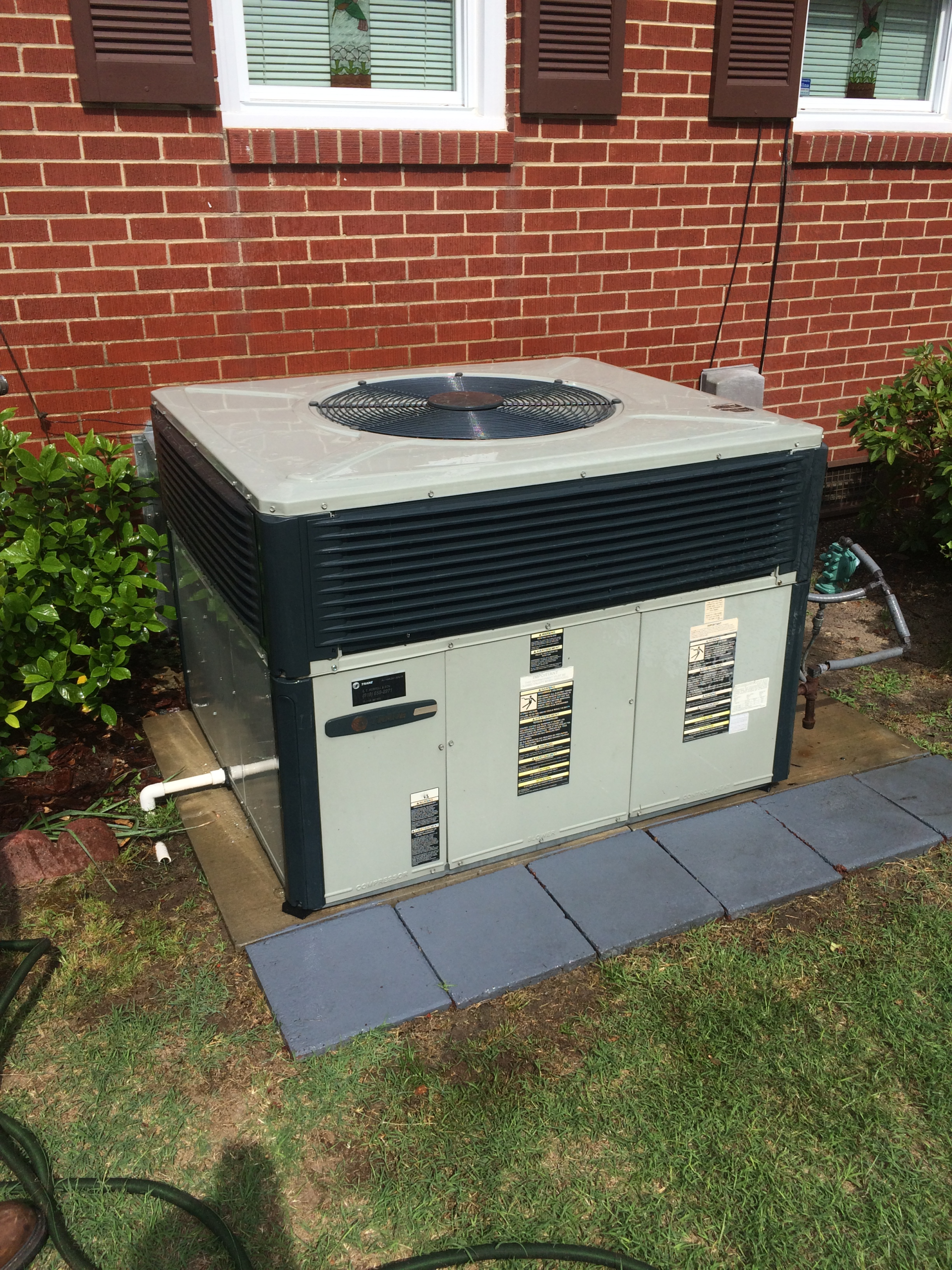 Air conditioning unit outside a brick building. Black top with side vents, gray panels, on gray square pads.