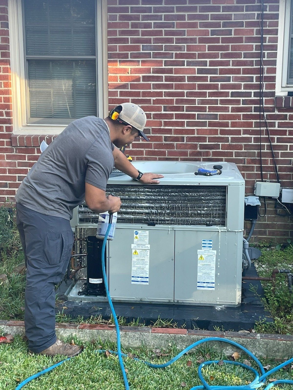 Person cleaning an air conditioning unit outside a brick building, wearing safety glasses and a hat.