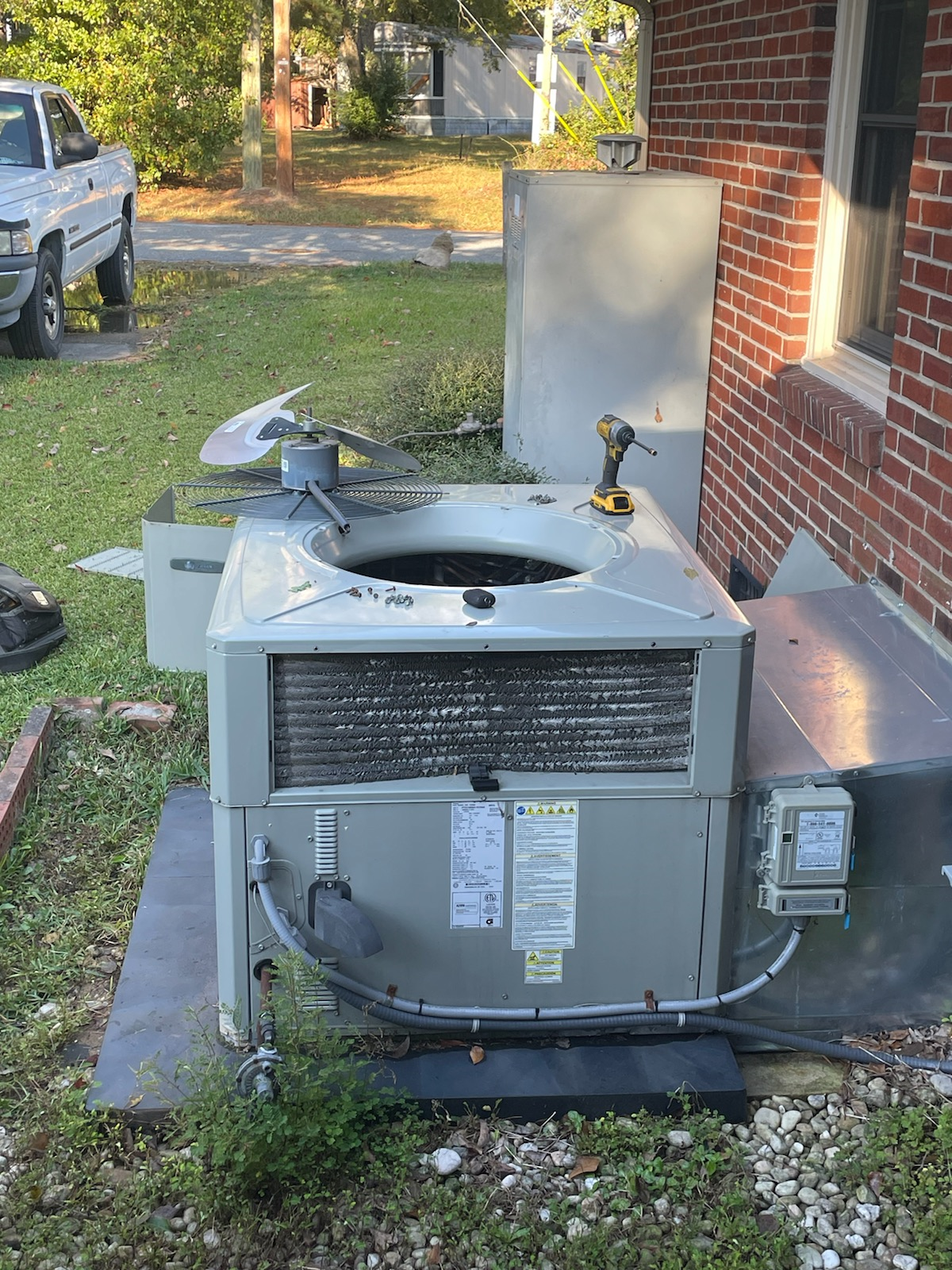 An air conditioning unit being serviced outdoors, near a brick building and a pickup truck.