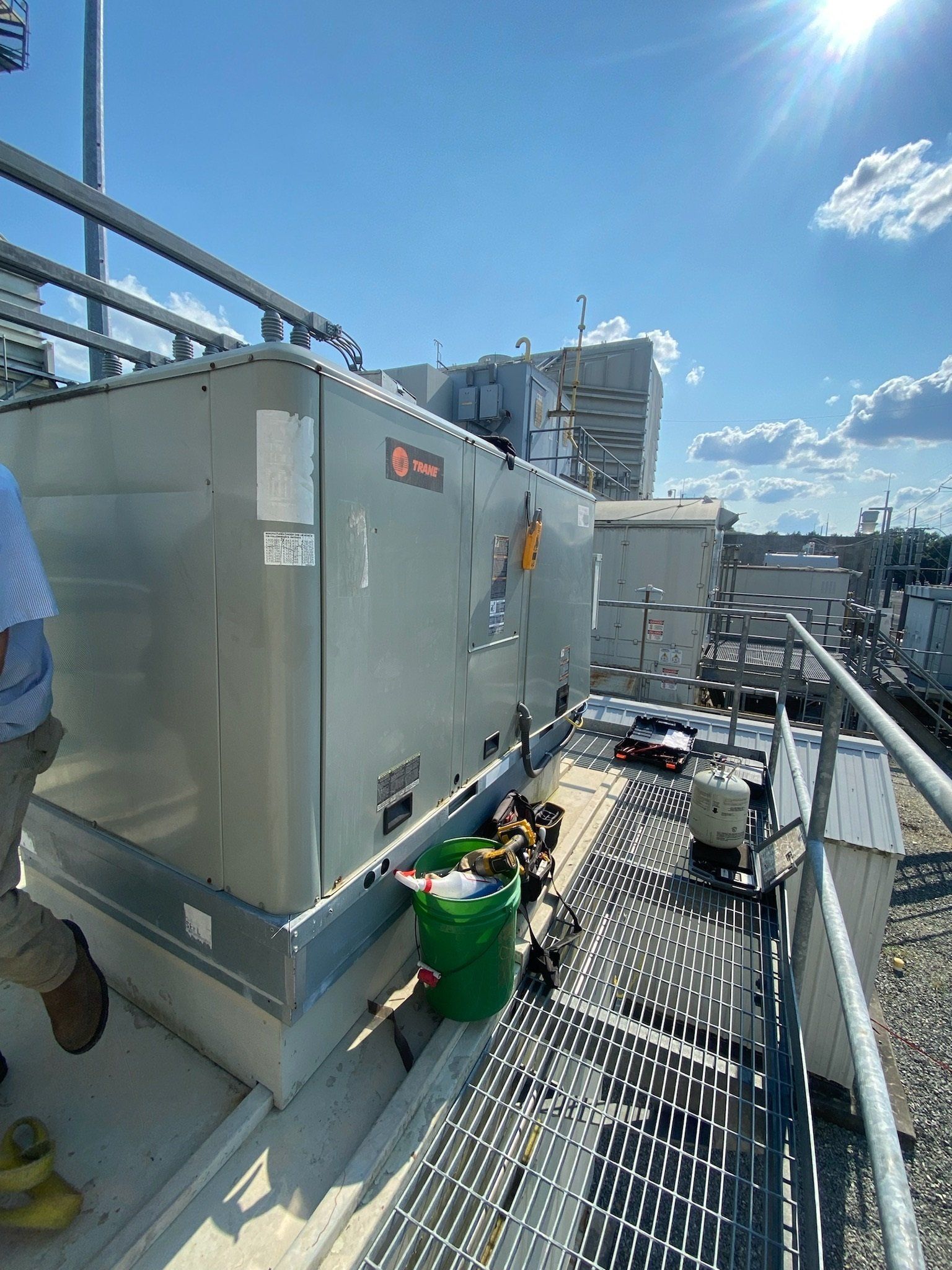 HVAC unit on a rooftop, with a green bucket and tools near it, sunny day.