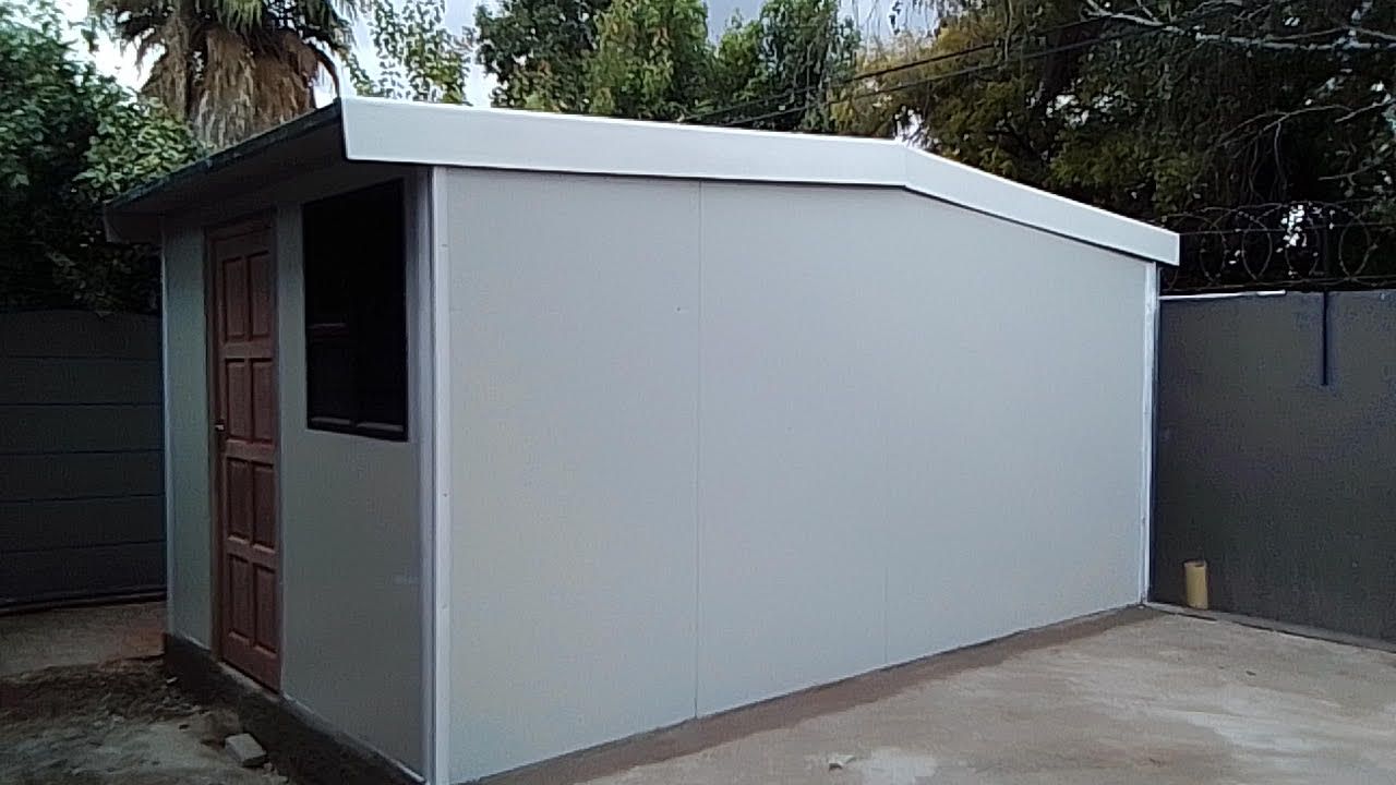 A small white building with a wooden door is in a backyard.