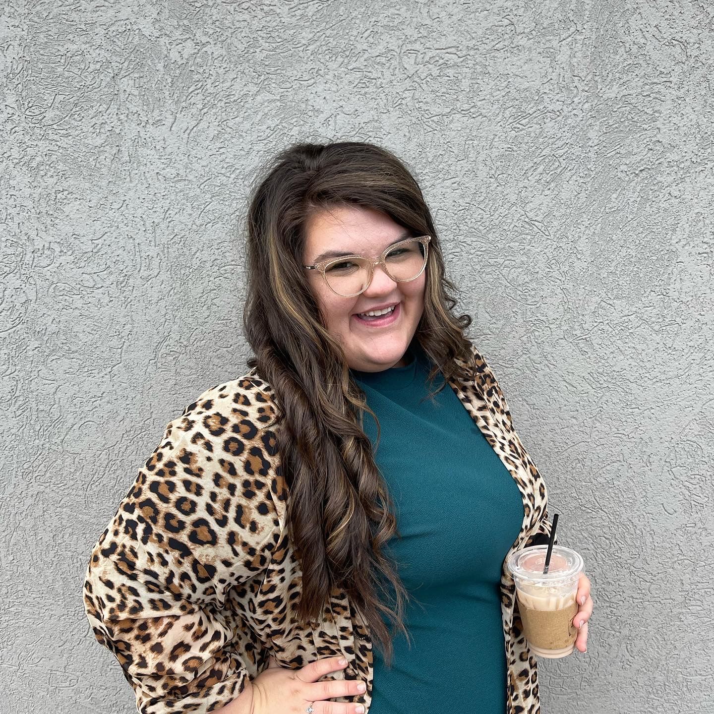 Smiling person in a leopard-print jacket holding an iced drink against a gray wall