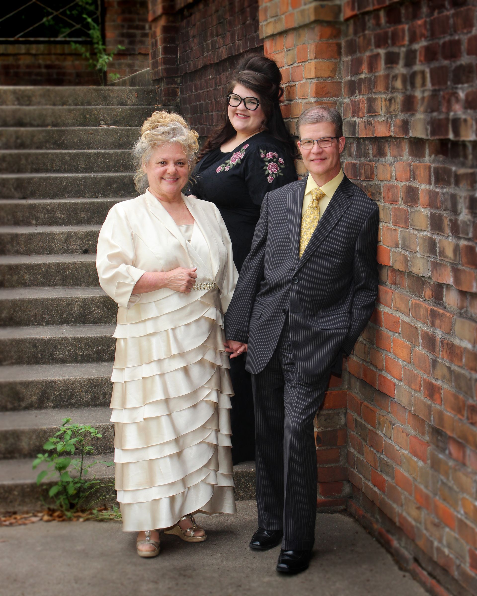 Three people pose near brick wall and stairs: woman in white dress, woman in black top, man in striped suit.