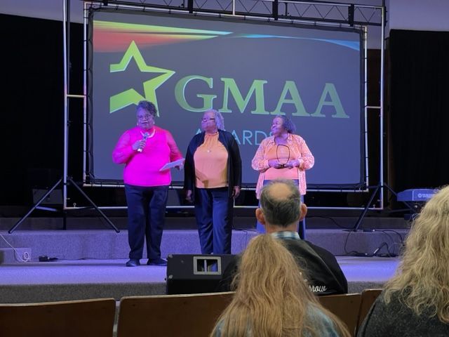 Three women stand on a stage in front of a screen that says gmaa