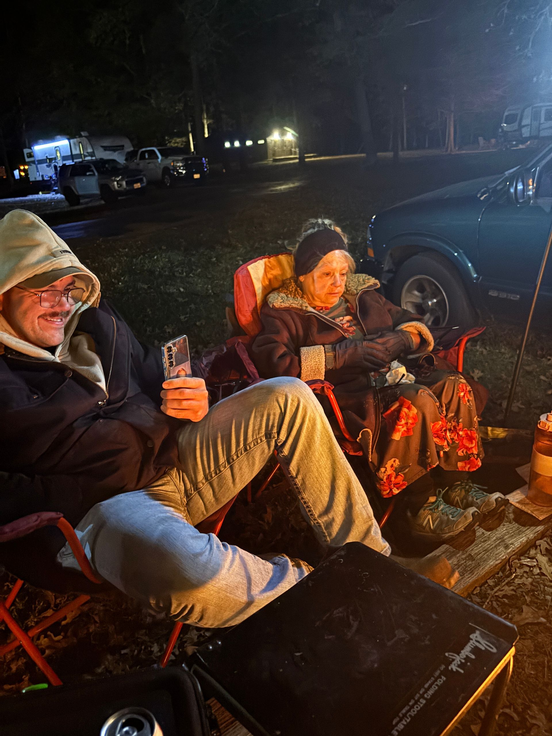 Two people seated in camping chairs at night. One holds a card, both bundled in warm clothing near a campfire.