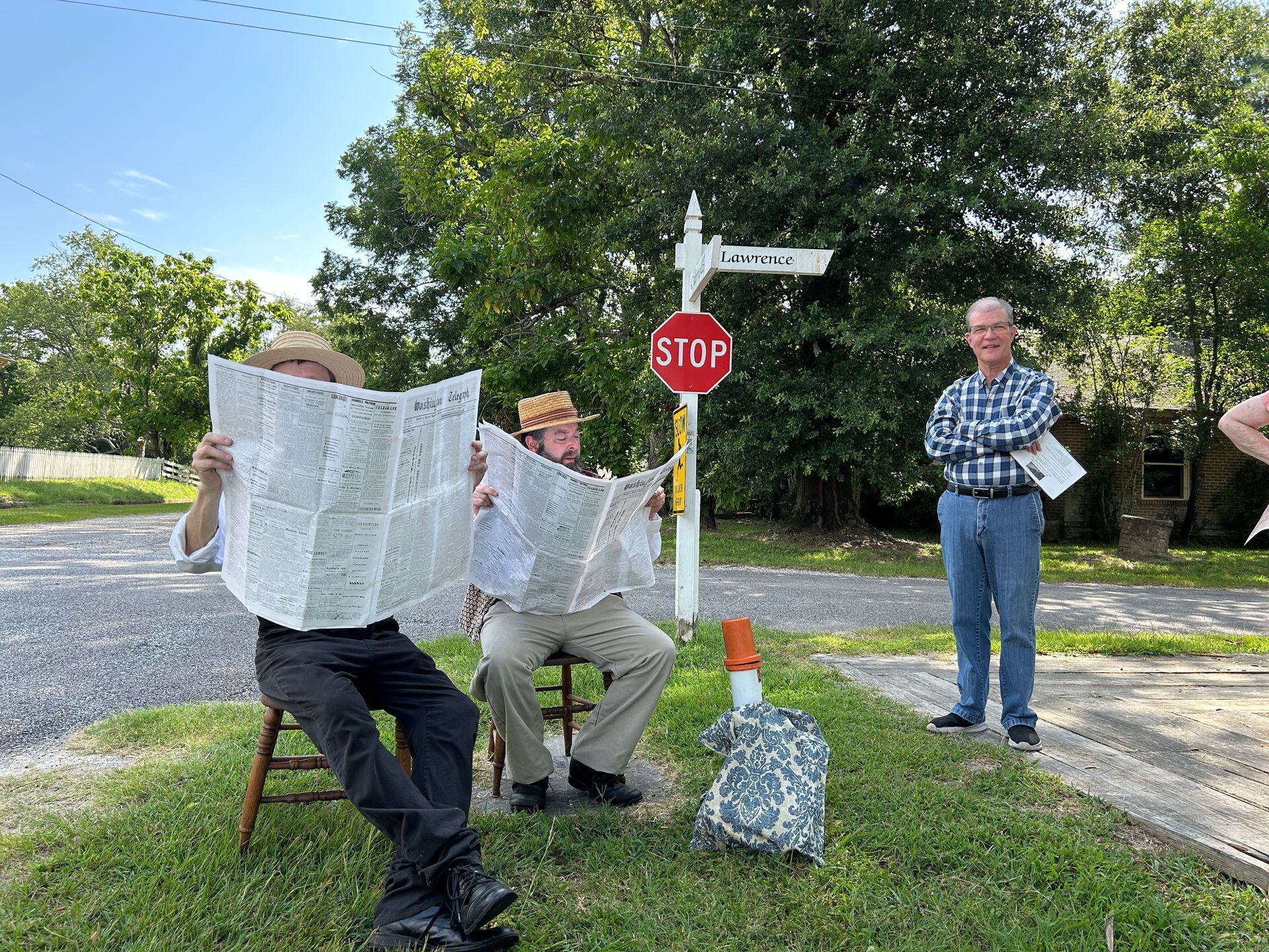 Two people sit reading newspapers by a stop sign while another stands nearby on a sunny street.