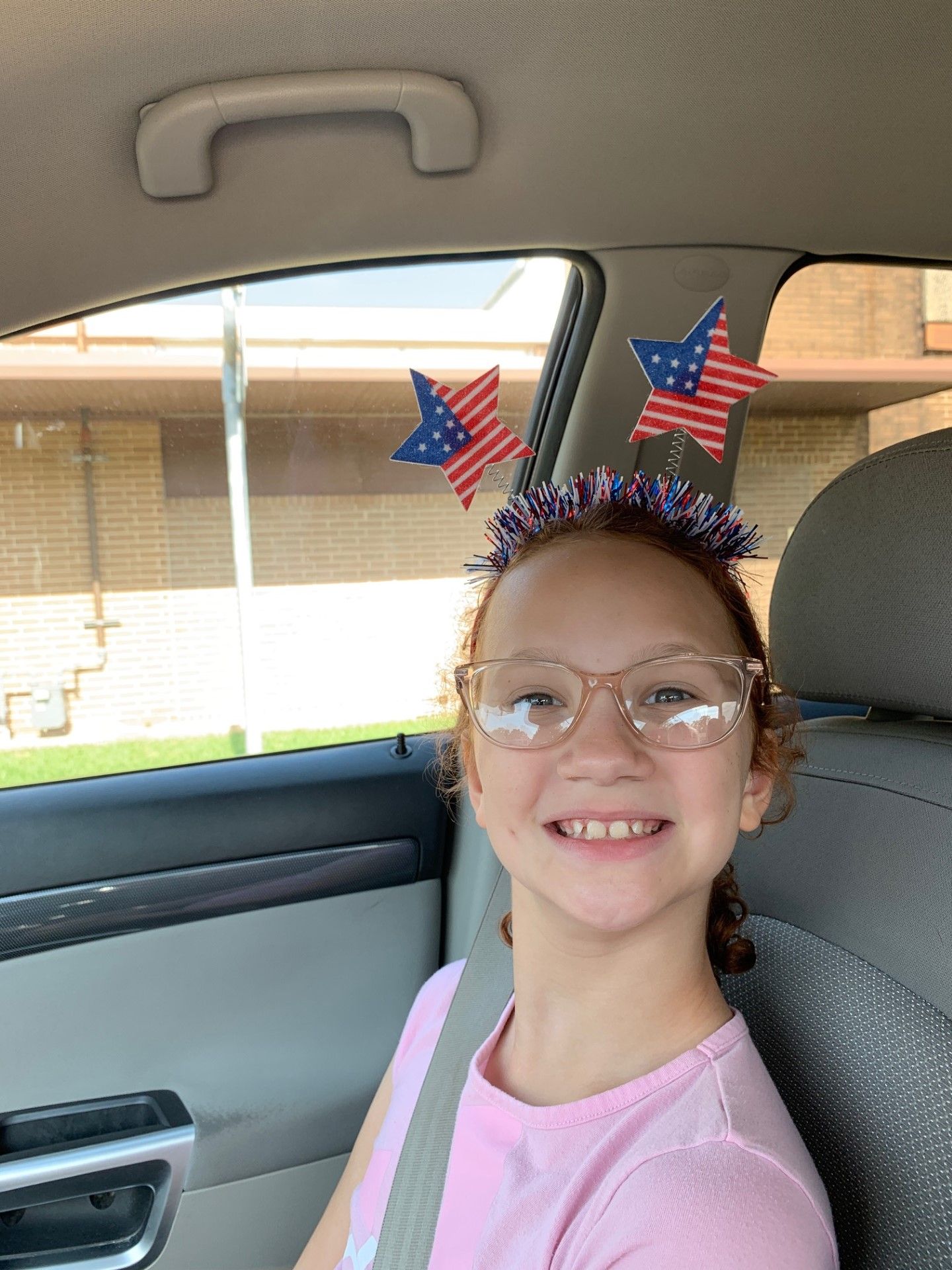 A young girl wearing glasses and a headband with american flags on it is sitting in a car.