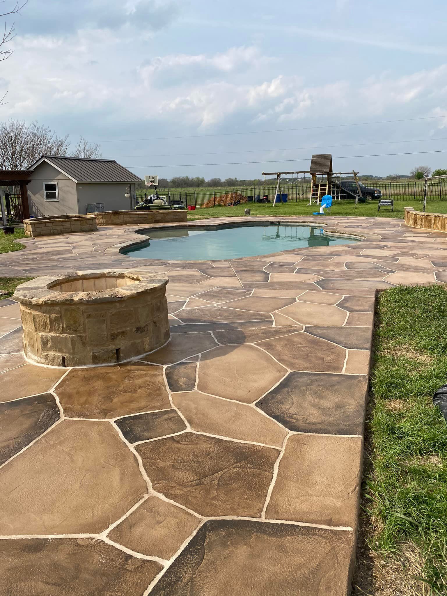 Stone patio with fire pit, pool, small building, and play set under cloudy sky.