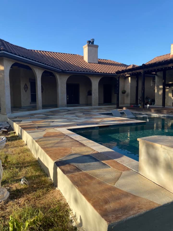 Patio and pool with stone coping; house with arches in the background; sunny day.