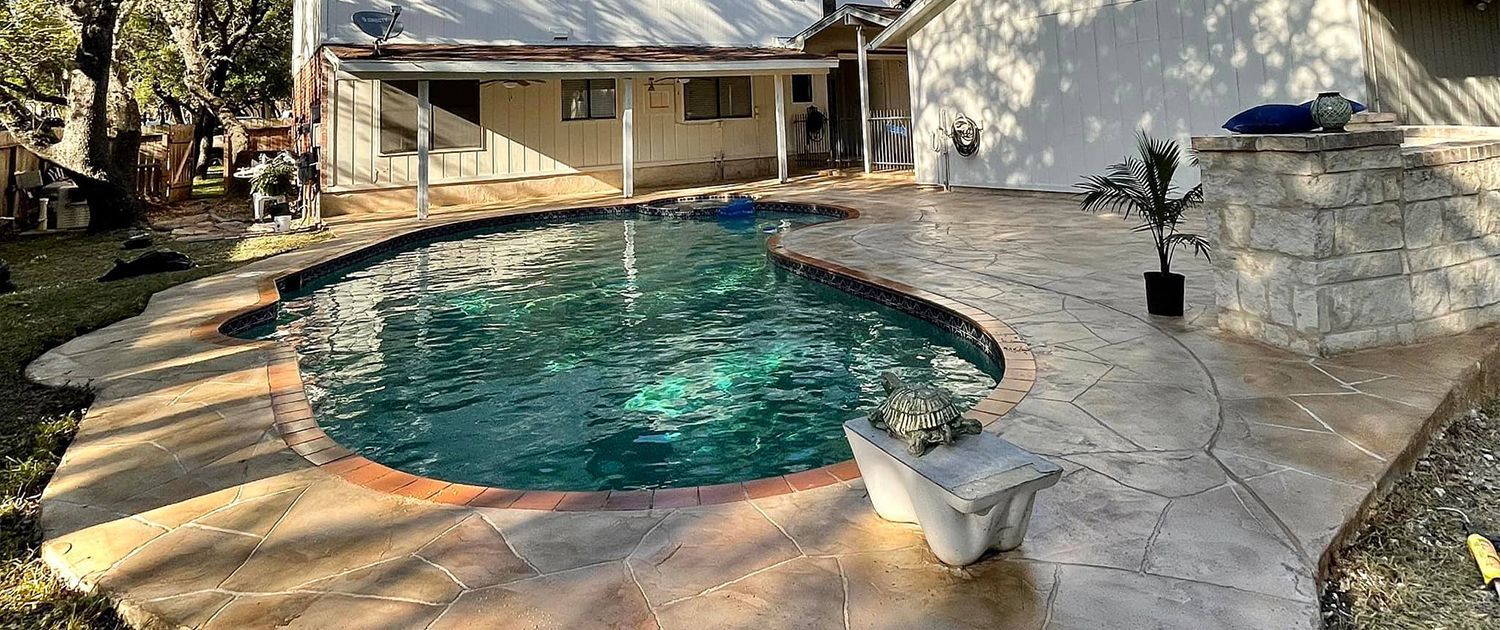 Backyard with pool, stone patio, and house. Sunlight casts shadows on the pool water and surroundings.